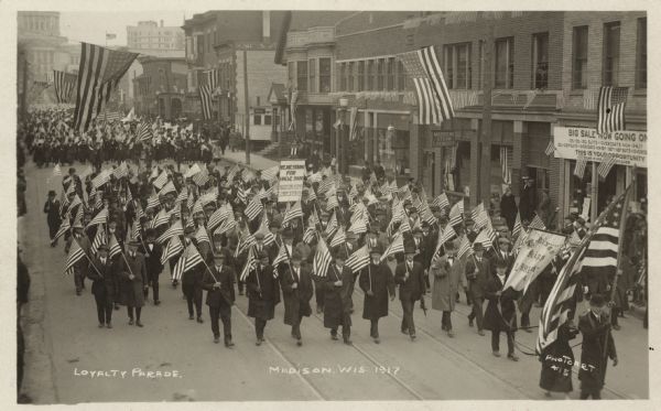 Elevated view of a parade marching down State Street. Large American flags hang from wires strung across the street. Madison Kipp employees, wearing suits and ties, are marching in a group and holding American flags and banners. The Wisconsin State Capitol is in the far background.