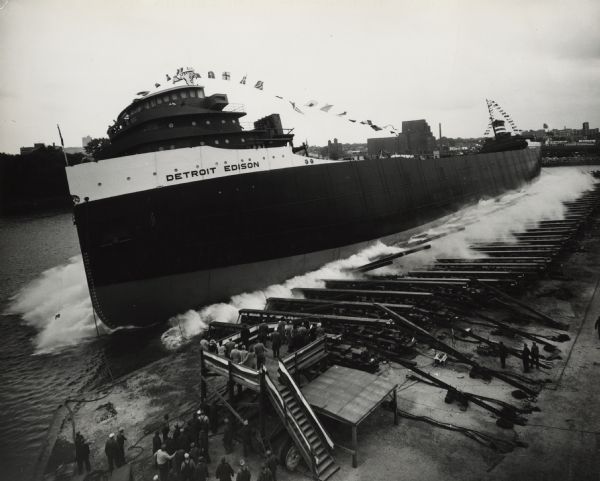 Elevated view of the sideways launching of <i>Hull 418, the Detroit Edison</i> at Manitowoc Shipbuilding, Inc. A group of people are gathered at the wharf on the lower left, and other people are on a platform near the slipway.