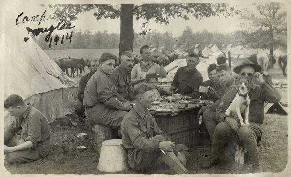 Group portrait of a number of soldiers, and a young boy, at Camp Douglas having a meal on an improvised table near a tree. One of the men has a dog on his lap. Tents are on the left and in the background. Horses are standing in the background on the left.