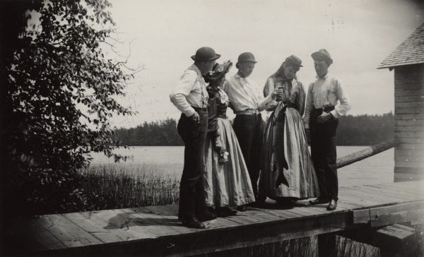 A group of three men and two women are standing on a pier inspecting a 15 pound muskellunge. Long Lake is in the background. There is a boathouse at the end of the pier on the right.