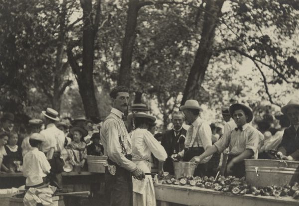 Exterior view of the punch table at a company picnic under trees. A man stands in the foreground looking at the camera while holding a knife, and the woman standing behind him is cutting up lemons. A man on the far right appears to be squeezing a lemon into one of the washtubs. On the tables are washtubs and many squeezed lemons. A large crowd of people are standing around the tables. Identified as the Milwaukee Electric Railway and Light Co. employees third annual outing at Waukesha Beach, Wisconsin.