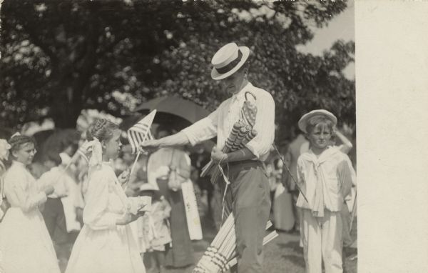 Postcard of a view of a man wearing a straw boater passing out flags to children on the Fourth of July. Text on reverse, "Sane 4th."