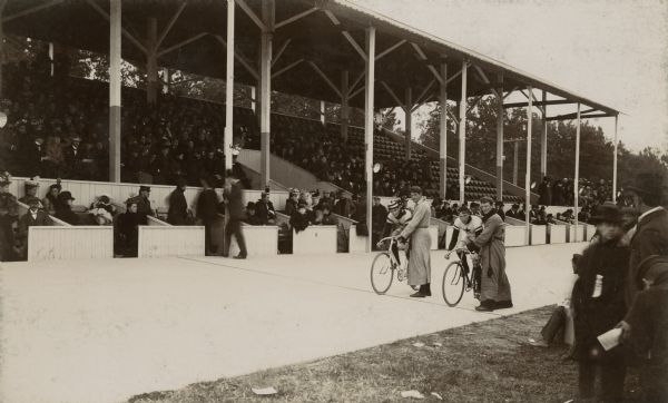 View from infield of two bicycle racers at the starting line of the quarter-mile cement bicycle track maintained by the Racine Athletic Association. Two men stand holding the two racers upright. The roofed grandstand on the other side of the track is crowded with spectators, and in front are viewing boxes.