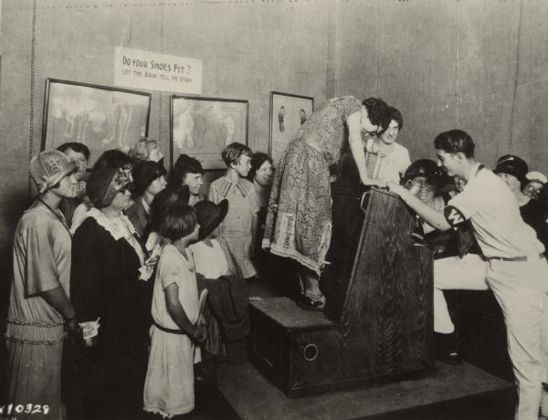 A group of women and children surround a woman who is standing on an X-Ray machine (fluoroscope) to determine if her shoes fit. A man on the right is operating the machine. A sign on the wall reads: "Do Your Shoes Fit? Let the Xray tell the story."