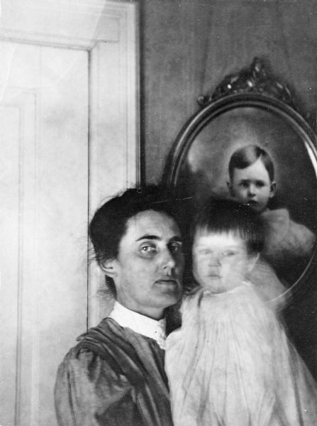 Interior portrait of a mother holding a child. On the wall behind them is a portrait of a child.