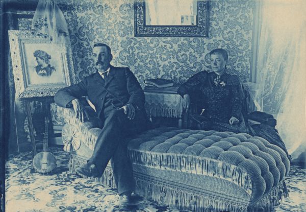 Interior portrait of Mr. and Mrs. Clint Smith. He is seated on a fainting couch on the left, and she is sitting in a chair behind him on the right. There is a painting of a woman on an easel on the far left, and a banjo is on the floor leaning against it.