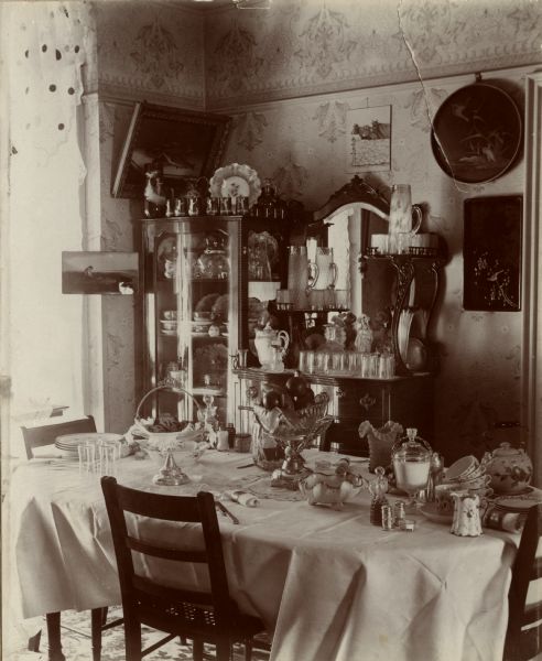 Interior view of the dining room in the James Nevins home on Broom Street. Dishes, teacups and glasses are stacked on the table. A bowl of fruit is in the center and on the left is a teacake stand with bread or cakes. A sideboard and china cabinet are against the far corner near a window.
