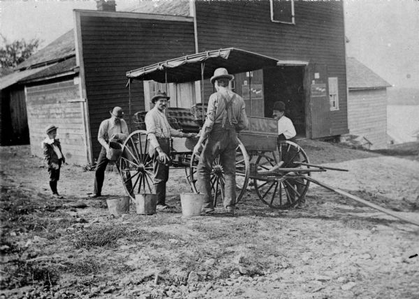 View across yard towards four men washing a surrey using metal buckets in front of a stable. A boy wearing a suit, necktie and straw hat is standing behind them on the right. Other buildings are down a hill behind the stable on the left.