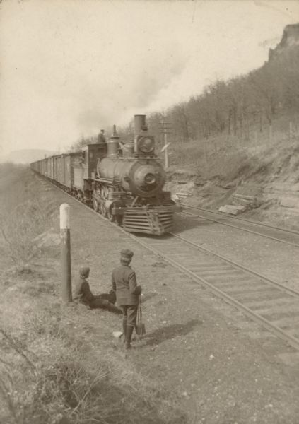 Slightly elevated view of two boys near a post at the side of two sets of railroad tracks. The boys are watching an oncoming train. On the right is a steep hill with a bluff at the top.