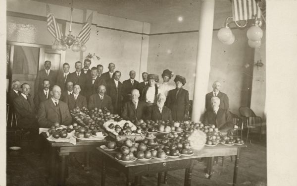 Interior group portrait of horticulture exhibitors and many plates of apples. The ceiling lamps are decorated with flags.