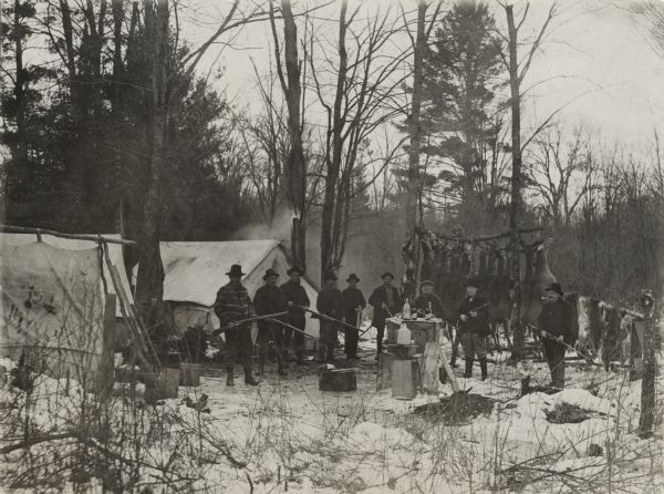 View of a hunting camp with the hunters assembled with their rifles in front of deer carcasses.