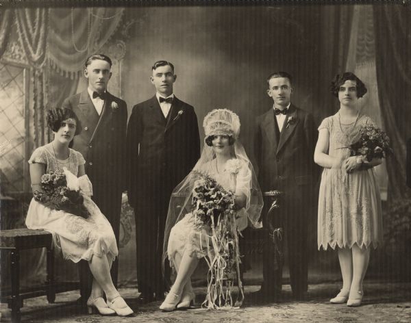 Studio group portrait of a wedding party. The bride, groom and three attendants. The women are holding bouquets. In the background is a painted backdrop.