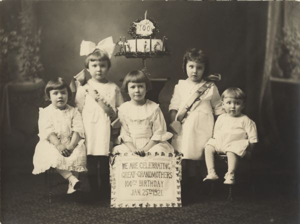 Studio portrait of five children posed in front of a cake, celebrating their great-grandmother's 100th birthday. The child in the center is holding a sign that reads: "We are celebrating Great-Grandmothers 100th Birthday Jan. 25th, 1921."