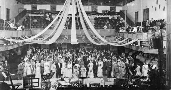 Elevated interior view of prom attendants assembled in a gym. Streamers are hanging from the ceiling. Some people are watching from the balcony.