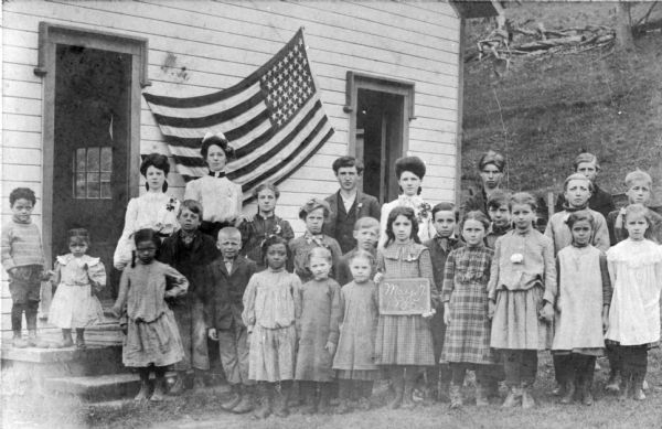 Outdoor group portrait of students and teachers of an integrated class at Eastman School. They are posing in front of the schoolhouse, and a flag is displayed behind them.