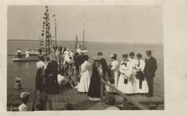 A group of men and women standing on a dock watching sailboats cast off. The tree covered shoreline is visible in the distance.