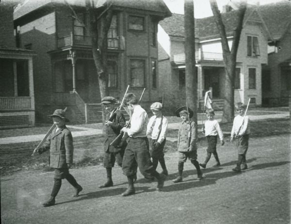 Boys marching down a neighborhood street with toy guns. The leader is carrying a big stick. A boy in the back is carrying a flag.