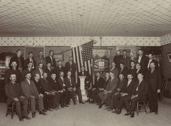 Interior group portrait of the United Brotherhood of Carpenters and Joiners of America, Local 1074 in Eau Claire, Wisconsin. Three rows of men with a flag and chapter banner in the center.