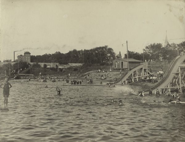 View across the water near the shoreline of the municipal swimming pool. The pool may have been along the Wisconsin River with slides and sandy beaches. A large group of bathers are playing in the water.