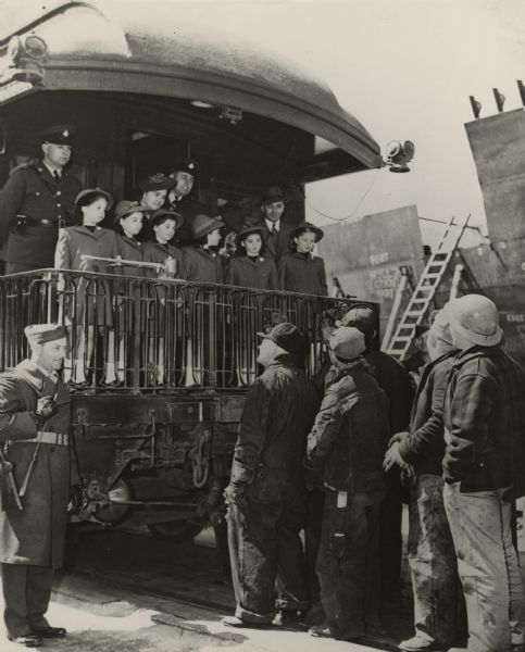 The five Dionne quintuplet girls, their sister and parents standing at the end of the observation car occupied by the Dionne party. They were in Superior to launch five cargo ships built by Walter Butler Shipbuilders, Inc., for the U.S. Maritime Commission. In the background on the right, a section of the hull of an escort can be seen, one of twelve being constructed in the Butler Shipyard. The quints are talking to shipyard workers.