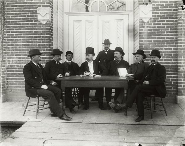 Outdoor group portrait of magistrates sitting around a table in front of the fire house. Identified (left to right): Edward Funke, William Schuster, David Thoeny, John L. Uttermuehl (with top hat), Chris Florin (standing), Gerhard Westerkamp, Marcus Fugina, J. Jacob Wunderlich.