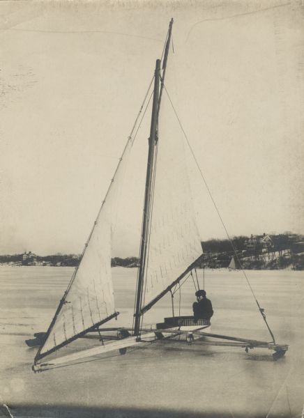 View across ice towards a man identified as Irving Brown sitting in an ice boat on a lake. Large houses are in the background along the shoreline.