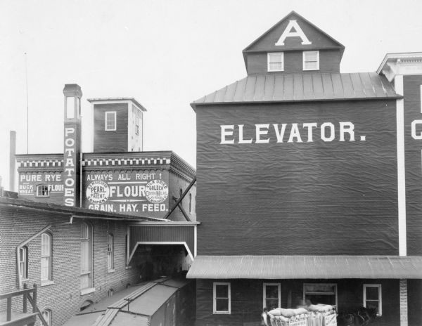 Slightly elevated view of the Cereal Mills Company. Men are working near a horse-drawn wagon near an entrance to a grain elevator in the lower right. Railroad cars are under an archway between the elevator and a brick building on the left.
