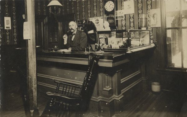 A man with a moustache stands smoking a cigar behind a hotel reservation desk. Behind him is a clock on the wall and a gramophone. A glass case on the counter on the right displays cigar boxes, and there is a rocking chair in the foreground.