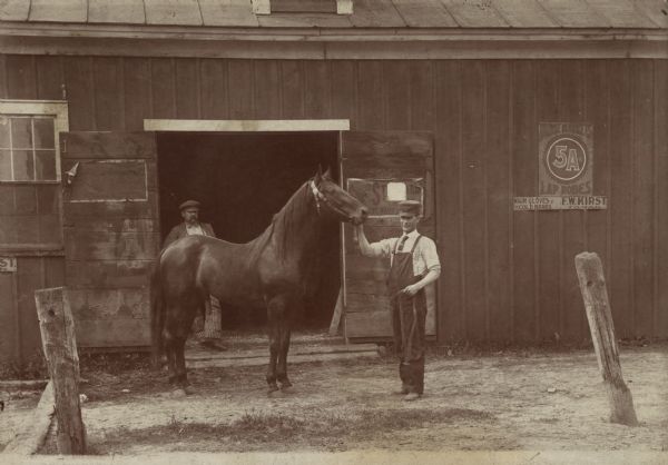 A man stands displaying a horse in front of the entrance to Frank Kirst's Harness Shop. A man is standing in the open doorway of the shop.