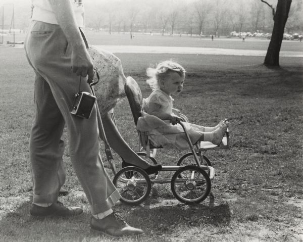 Man, seen from waist down, pushing a small girl in a stroller in Vilas Park. He is carrying a camera in his right hand.