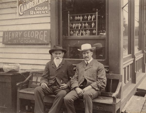 Carl Bohnsack (left) and August Marquardt sitting on a bench outside a jewelry store. They are wearing hats and suits. Pocket watches are on display in the window and signs on the outside wall advertise cough syrup and cigars.