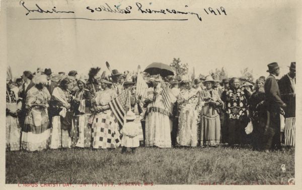A gathering of Native American women, some holding flags, to welcome home soldiers returning from WWI on Corpus Christi Day. There are men in the background. Handwritten on top, "Indian soldiers' homecoming 1919." Text in red on foot, "Corpus Christi Day, June 19, 1919, Reserve, Wis. Photo by N. F. Huie, 400 7th St. Racine, Wis."