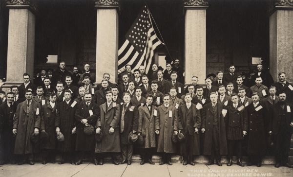 Group of WWI draftees assembled in front of the Ozaukee County Courthouse. In the background a man is holding up a flag between the center columns of the building. Text at foot of photo, "Third call of selectmen, Local Board, Ozaukee, Wis."