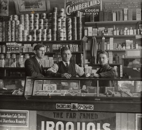 Three men posing behind the counter of a grocery store. Canned goods and jars of candy on the shelves behind them on the left, and pharmaceuticals are on the shelves on the right. Cigars are in the glass cases at the front of the counter.