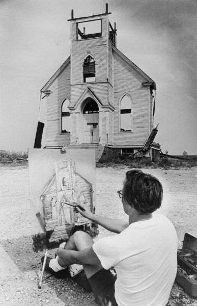 View of the front wall left standing of the Trinity Lutheran Church. A man in the foreground us painting a portrait of the church. The artist is identified as Peter Zirnis.