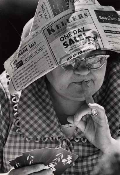 Close-up of a woman wearing a newspaper hat playing cards. Identified as Esther Lewis playing with the Golden Agers club members in Washington Park, Milwaukee.