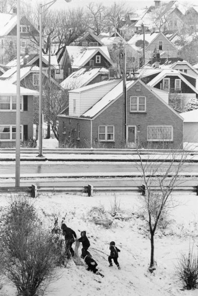 Elevated view of children sliding on the snow near the Bay Street overpass to Jones Island. The children are walking up the hill. In the background is a street with guard rails and beyond is a neighborhood.