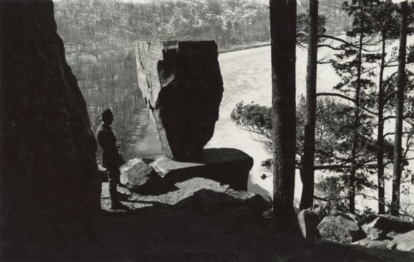 Elevated view of a man standing on a rock looking over a frozen Devil's Lake in front of a Balanced Rock. Original caption: "There are some startling rock formations on the bluffs surrounding Devil's Lake."