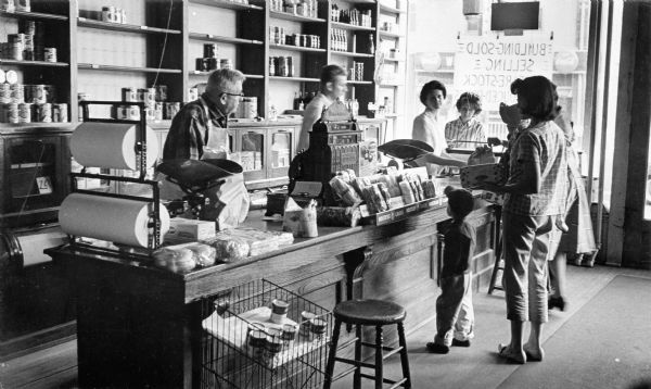 William Behnke selling out after 60 years as the neighborhood grocer at North Avenue and 29th Street. There are four people behind the counter, and customers standing in front. A sign on the window in the background reads: (backwards) "Building-Sold."