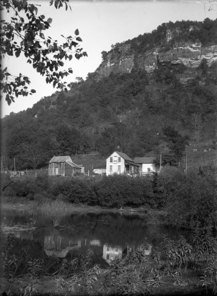 View across water towards a house on the banks of the Mississippi River at the base of rocky bluffs. Probably located between Alma and Nelson, along Beef Slough.