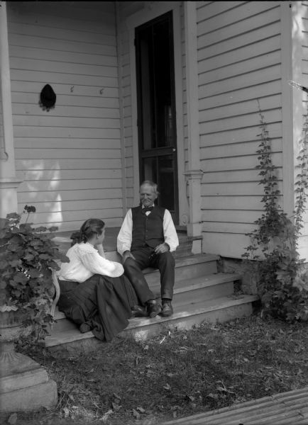 A man and a woman sitting on the front steps of a residence. Originally identified as Bertha Gesell and Uncle Christ, but later identified by Peter Gesell as his aunt Wilma and his great grandfather's brother.