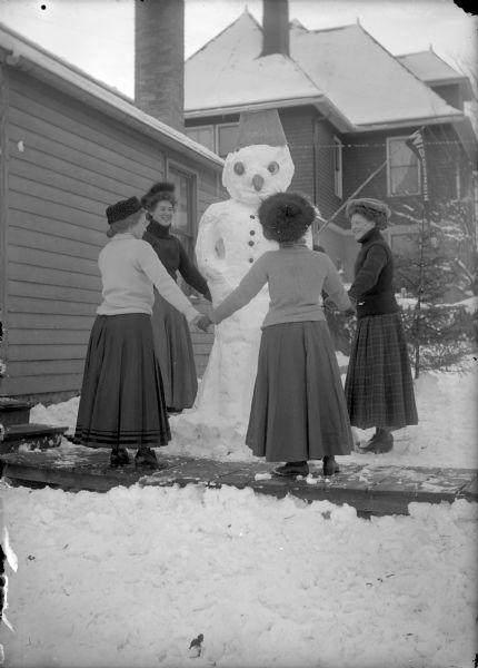 A group of women are holding hands in a circle around a snowman. The snowman is holding a UW pennant. Identified as: Vera Verhusen (back, left), Mary and Elsie Thom (right)