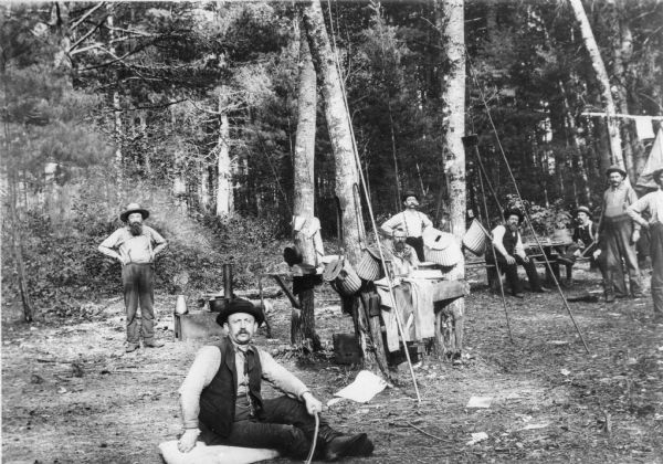 Group portrait of men at a fishing camp in the woods near Thunder Lake. Creels are hanging near long bamboo fishing poles leaning against trees.