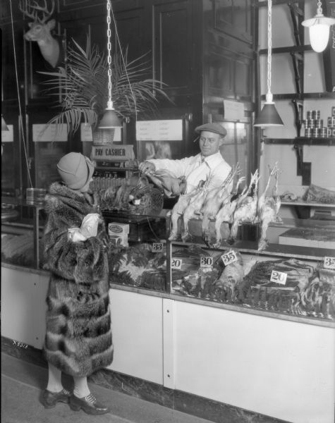 View towards a woman standing at the meat counter of the Madison Packing Company. She is buying chickens from a man standing behind the counter which has a display of meat in glass cases.