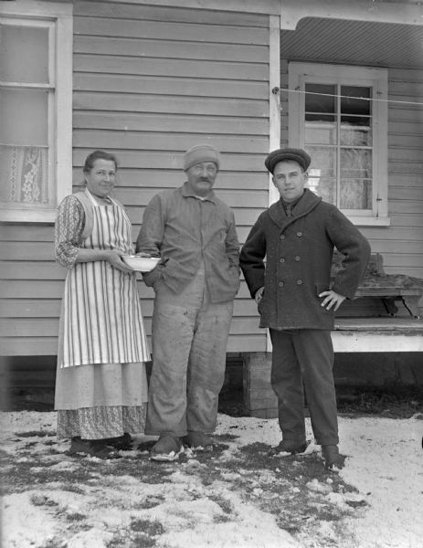 Man and a farm couple standing next to a house. The woman is holding a bowl and there is snow in the yard.