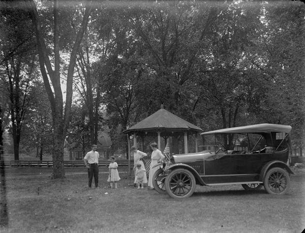 A group of a man, two women and two children playing croquet next to an automobile and a gazebo in a park.