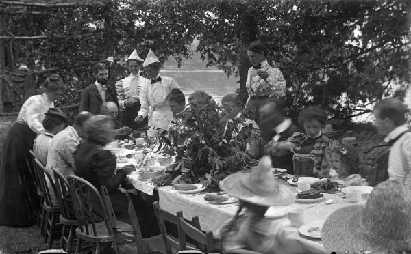 The Meinhardt family assembled for a picnic in their garden. A river or lake is in the far background. The table is set with china and flatware. Among the food served is corn on the cob and coffee.