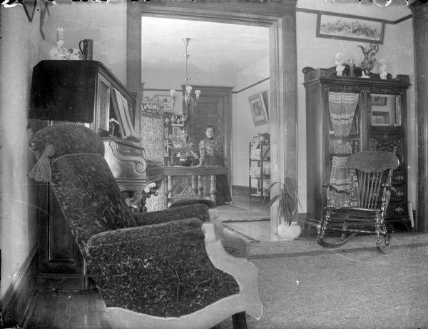 Interior view of the parlor and dining room of the Straubel home. A piano is in the parlor in the foreground, and a woman is standing next to the dining room table in the background.