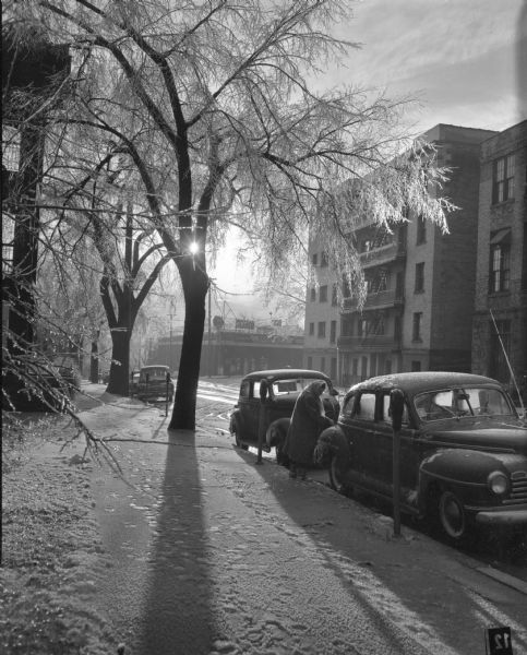 View of the 110 block of West Doty Street after a sleet storm. Cars are parked at the meters on the curb and a woman stands next to a car. The Baskerville Apartments are on the right. A Texaco Station is located on the corner in the background.