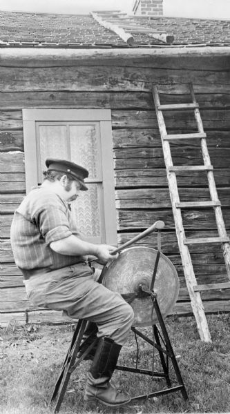 Man sharpening an axe on a grinding wheel next to a cabin at Old World Wisconsin.
